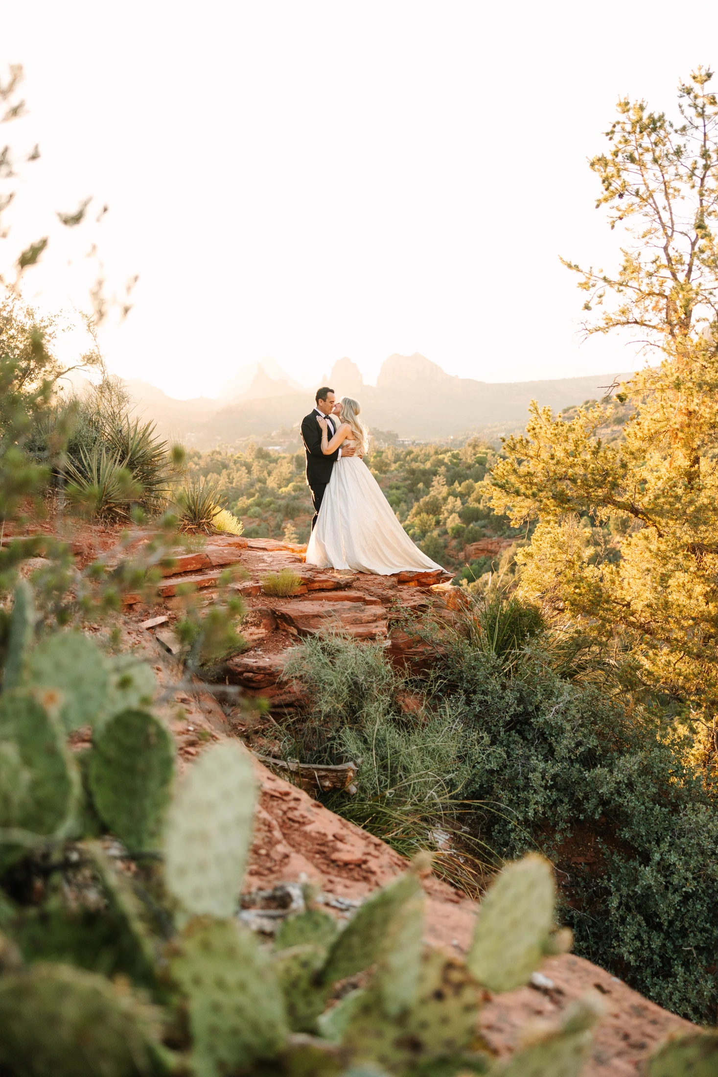 Shana and Kamal standing together among sunlit red rocks and greenery near the Munds Wagon trailhead