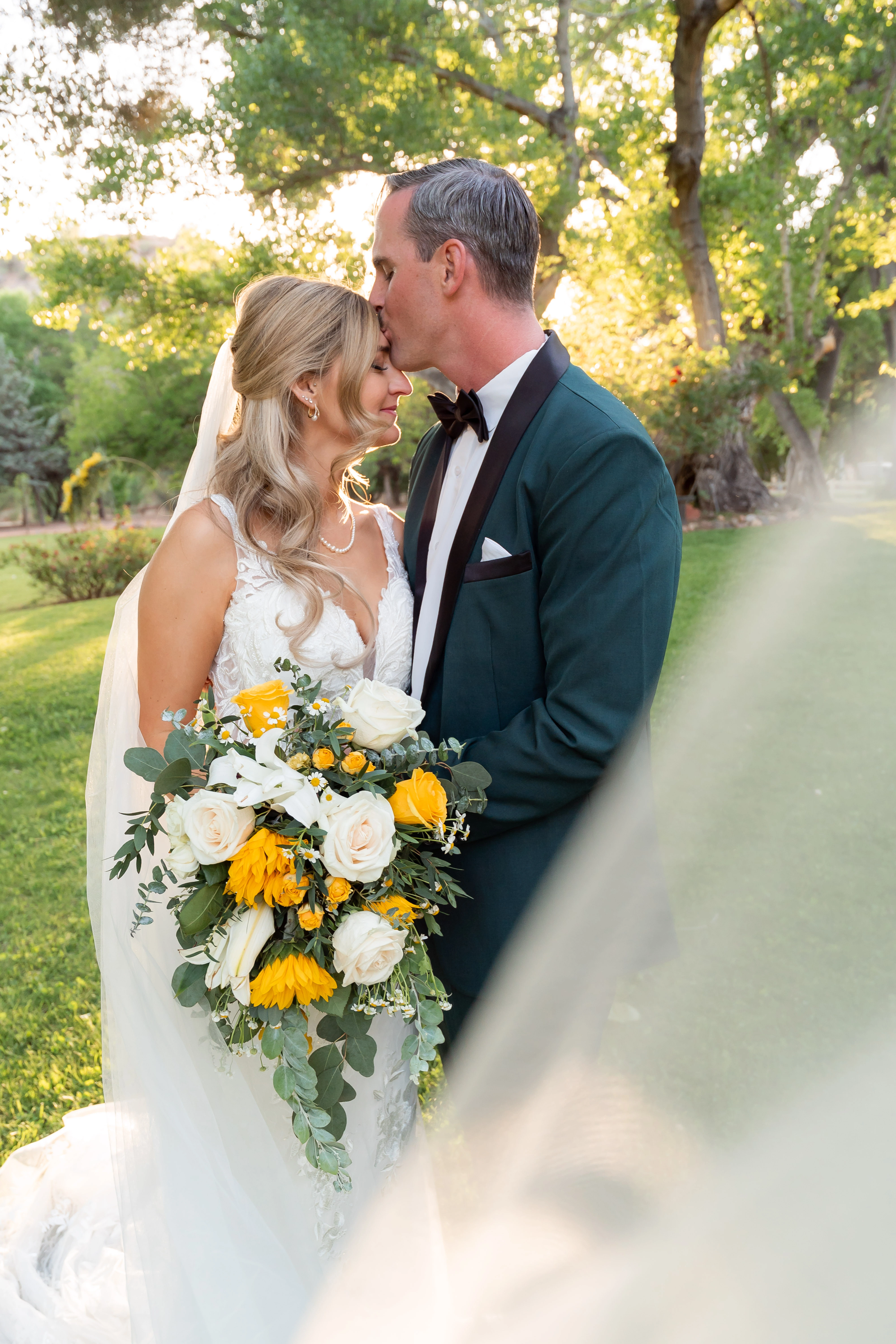Clayton kissing Tyler’s forehead beside her bouquet and sunlit veil at their Page Springs wedding